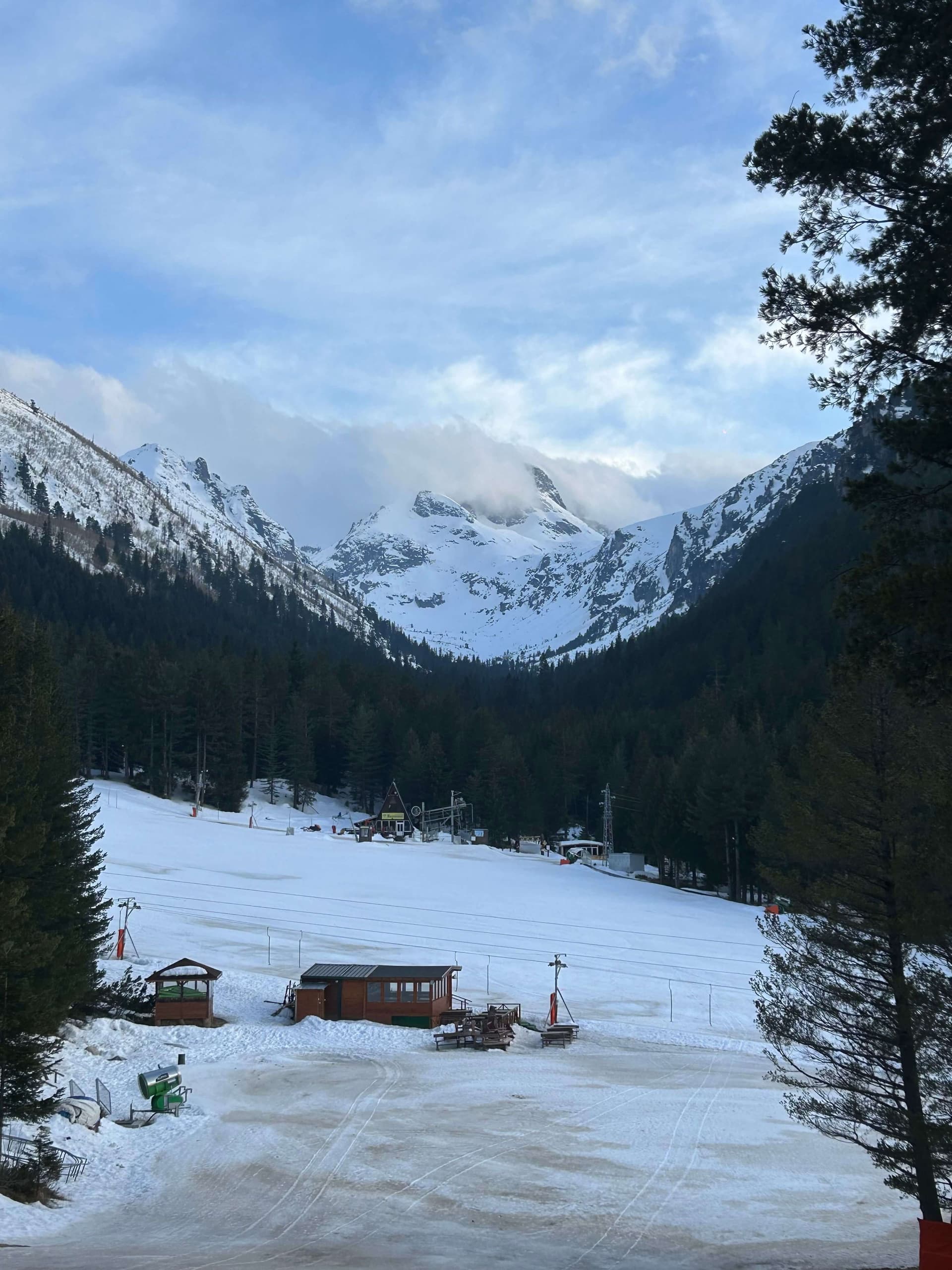 A wide, gentle beginner slope with drag lifts and wooden huts below the snowy peaks of Pirin