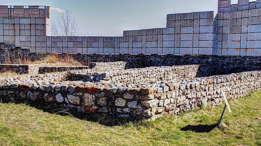 Excavated stone wall foundations at Krakra Fortress, with a reconstructed rampart rising behind them